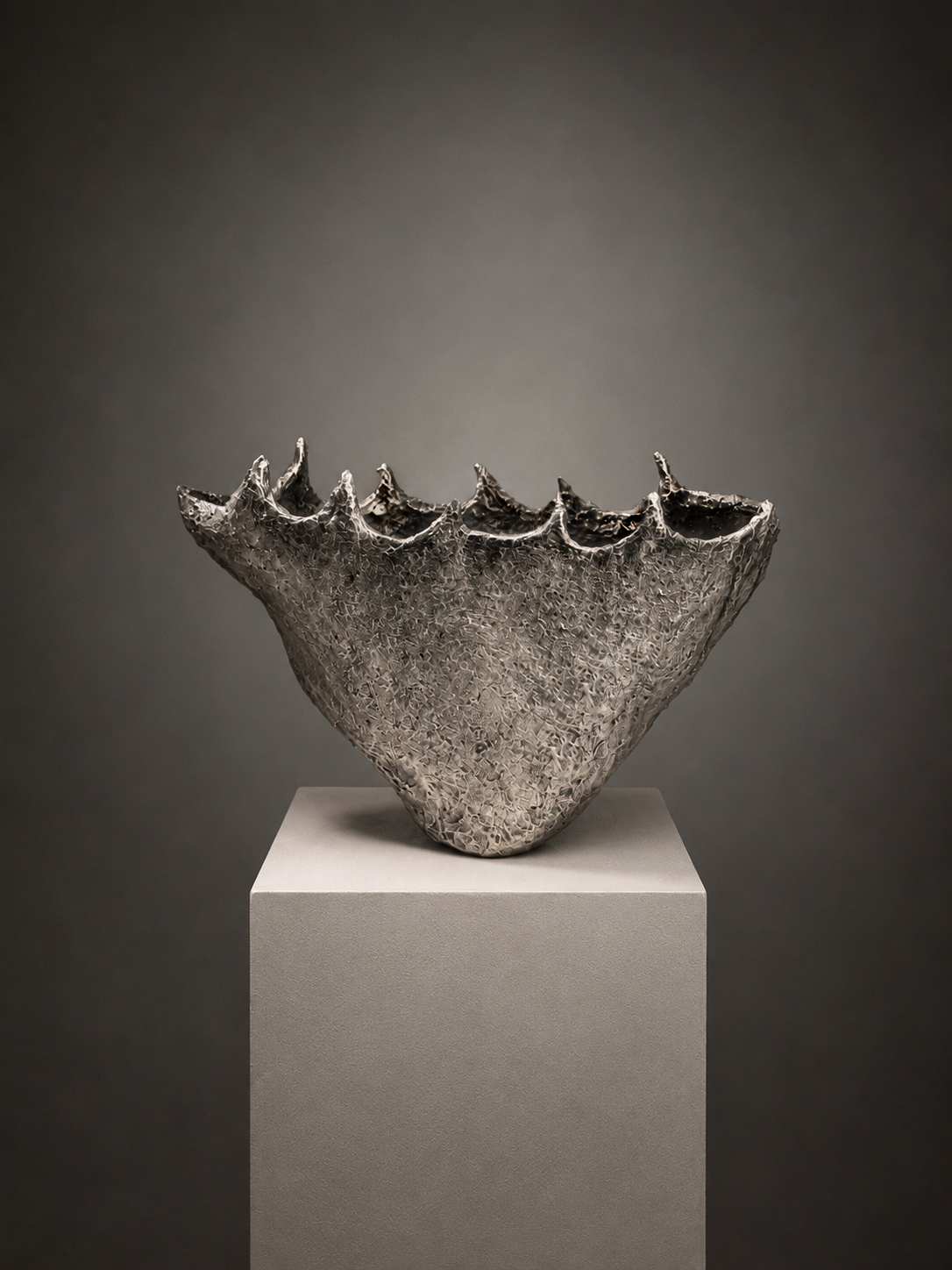 A sculptural metallic bowl with a textured, ribbed surface and irregular scalloped rim, displayed on a light gray pedestal against a dark gray gradient background.